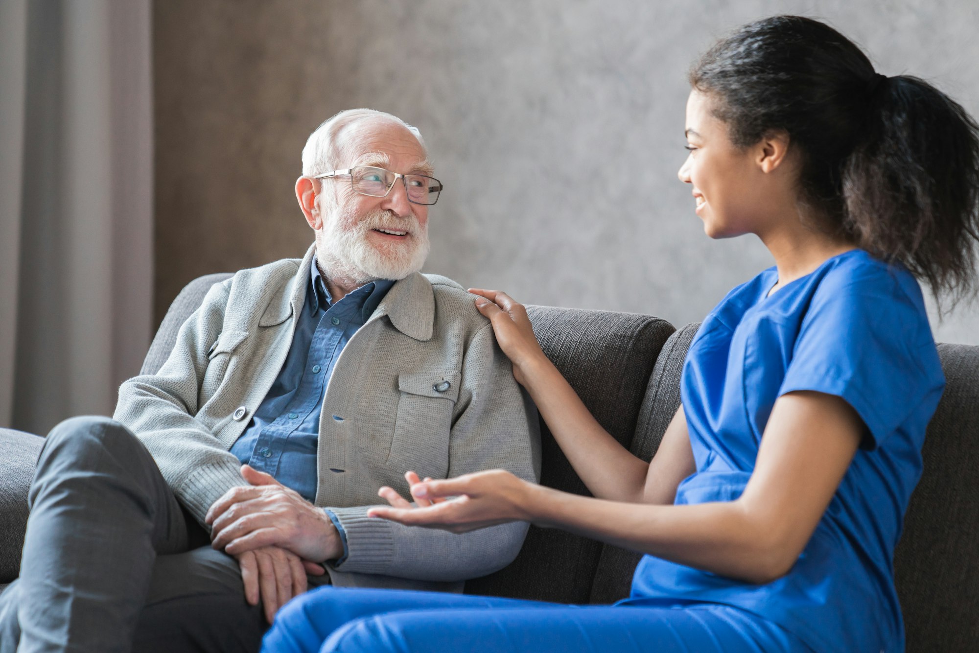Caring african-american nurse talks to old elderly senior patient holds his hand sit in living room